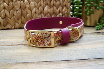 A dog collar with a floral patterned exterior, a burgundy accent strap, and gold-tone hardware, displayed on a rustic wooden surface with a woven basket and green plant in the background.