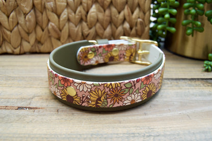 A dog collar with a floral patterned exterior, an olive green accent strap, and gold-tone hardware, displayed on a rustic wooden surface with a woven basket and green plant in the background.