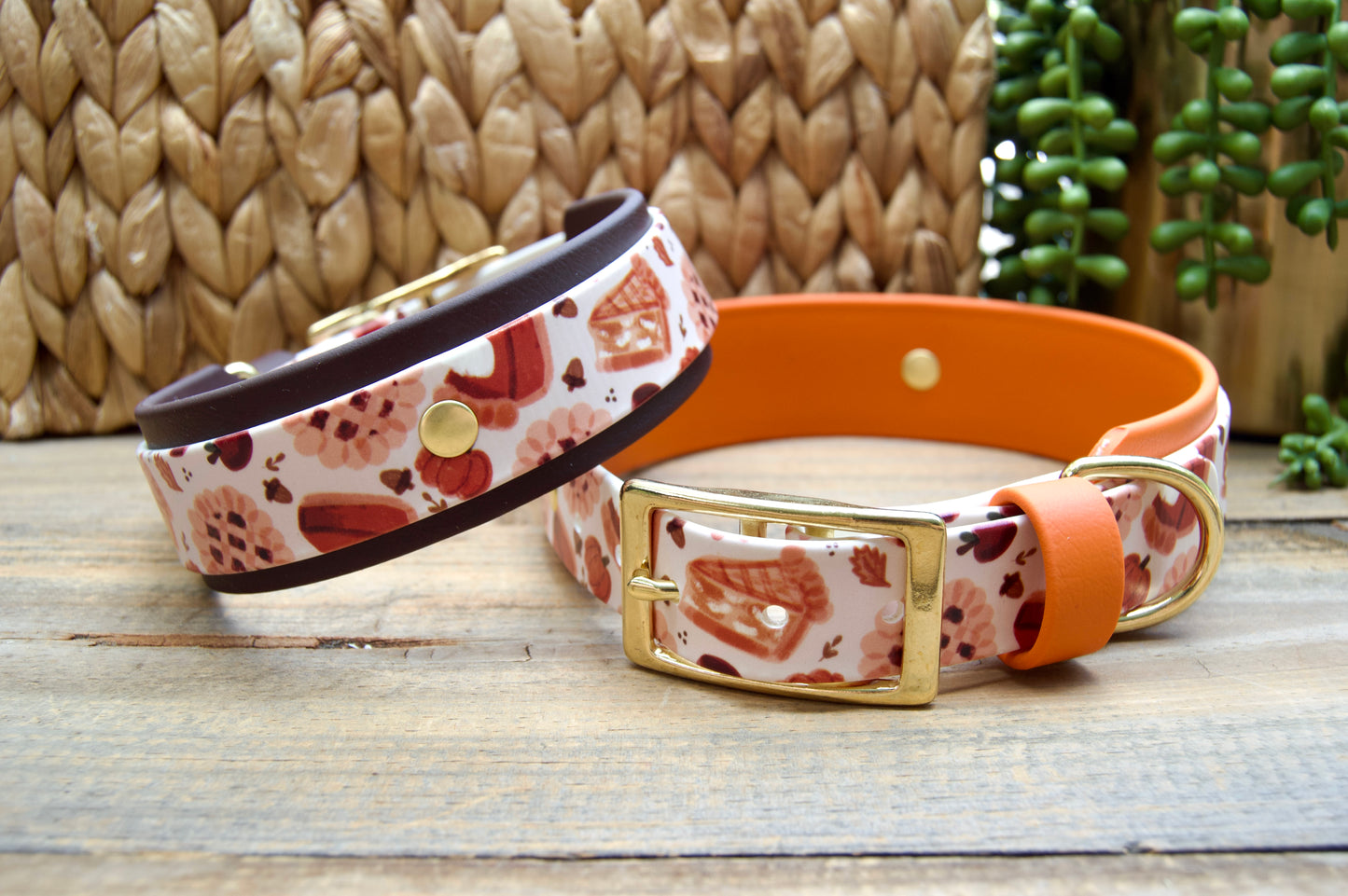Two dog collars displayed on a rustic wooden surface. The collar has a white base with a red and orange pie pattern and a dark brown accent color, while the other has a burnt orange biothane accent. Both have gold hardware.