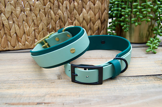 Two dog collars, Sage and Emerald Green Colors on a wooden surface with a woven basket and plant in the background.