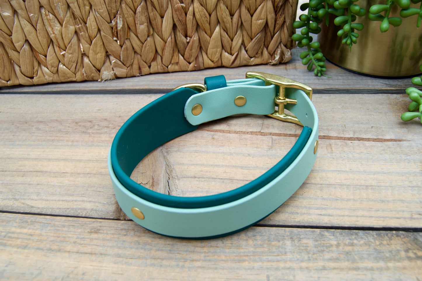 Sage and Emerald Green Colored dog collar on a wooden surface with a woven basket and plant in the background.