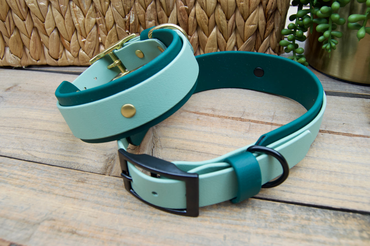 Two dog collars, Sage and Emerald Green Colors on a wooden surface with a woven basket and plant in the background.