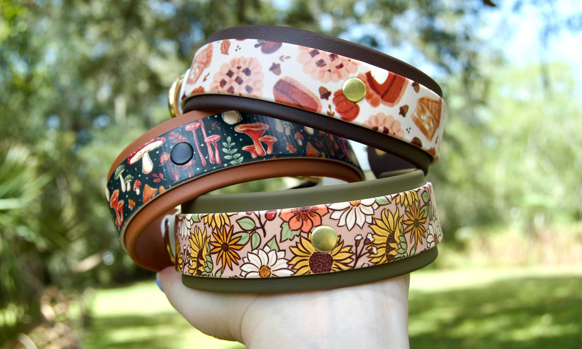 Three patterned dog collars stacked on a hand, photographed outdoors with a blurred green garden background. The collars are 'Fall Floral', 'Mushroom', and 'Pie'. 