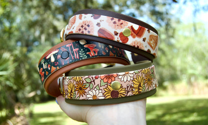 Three patterned dog collars stacked on a hand, photographed outdoors with a blurred green garden background. The collars are 'Fall Floral', 'Mushroom', and 'Pie'. 