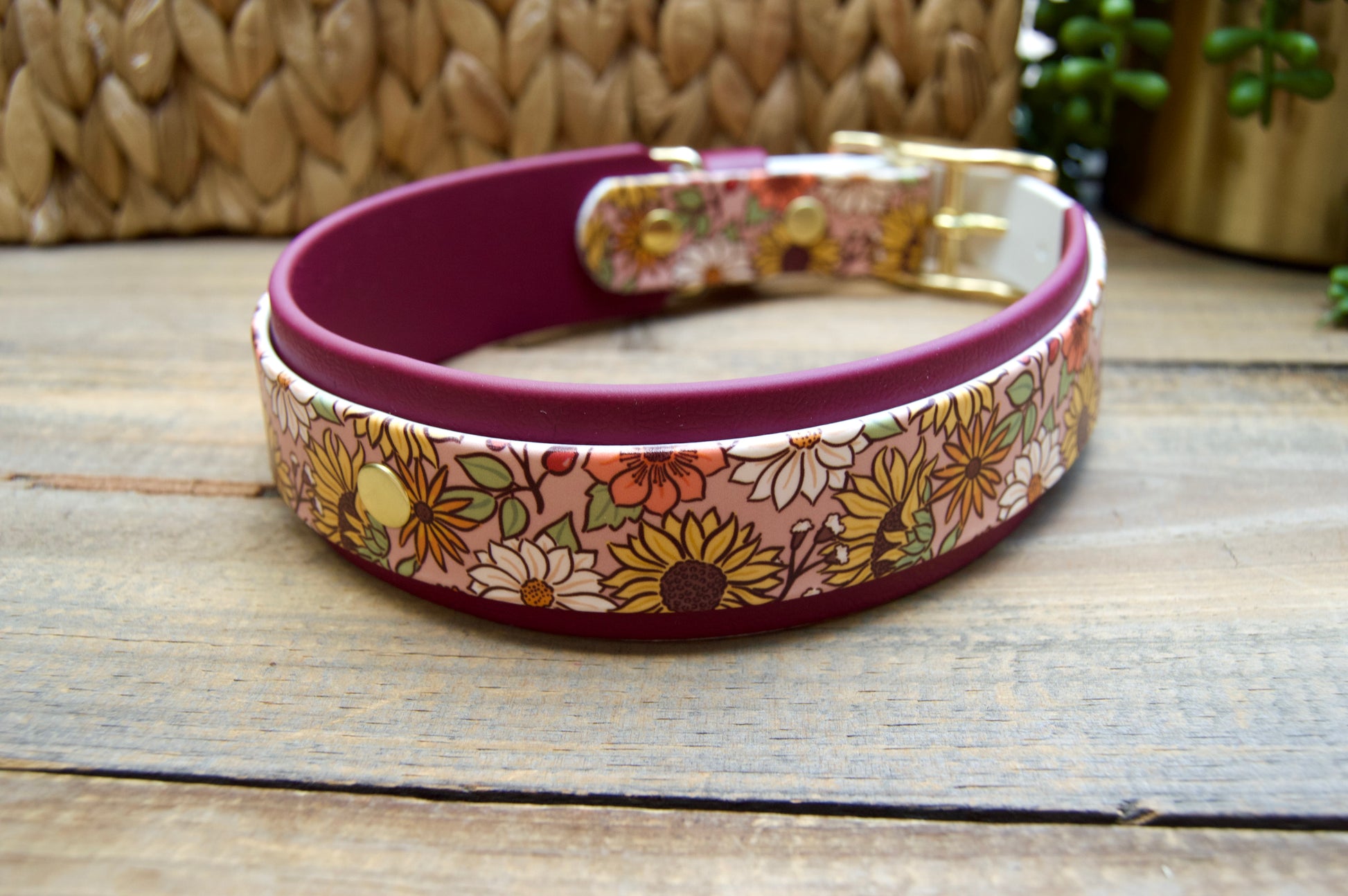 A dog collar with a floral patterned exterior, a burgundy accent strap, and gold-tone hardware, displayed on a rustic wooden surface with a woven basket and green plant in the background.