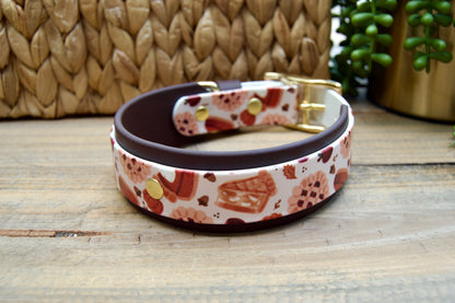 A dog collar displayed on a rustic wooden surface. The collar has a white base with a red and orange pie pattern and a dark brown biothane accent color with gold hardware.